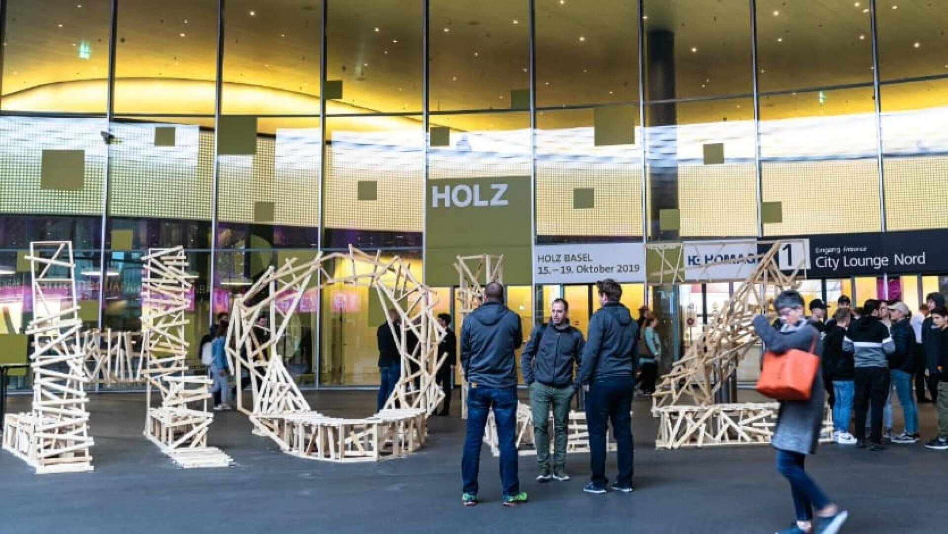 Menschen stehen und unterhalten sich vor einem modernen Glasgebäude mit der Aufschrift HOLZ. Draußen sind hölzerne Kunstwerke ausgestellt, und es sind mehrere Veranstaltungsbanner und eine Menschenmenge zu sehen.
