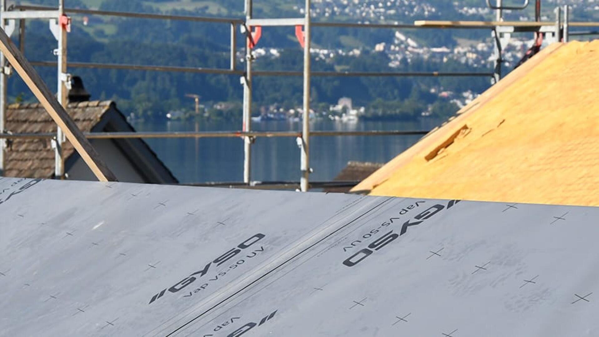 Chantier de construction avec, au premier plan, des échafaudages et un toit recouvert d'une bâche de protection. Un lac et des bâtiments sont visibles à l'arrière-plan, partiellement flous.
