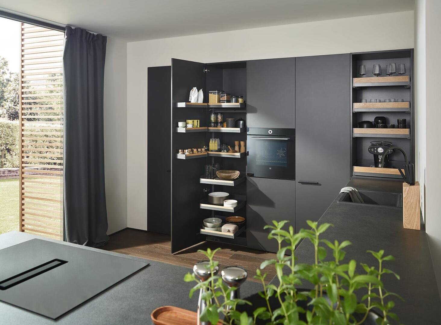 Eine moderne Küche mit mattschwarzen Schränken, einer offenen Speisekammer, die ordentlich geordnete Regale mit Lebensmitteln und Geschirr zeigt, einem eingebauten Backofen, Holzregalen mit Tassen und einer Kaffeemaschine und einer Topfpflanze auf der Arbeitsplatte.