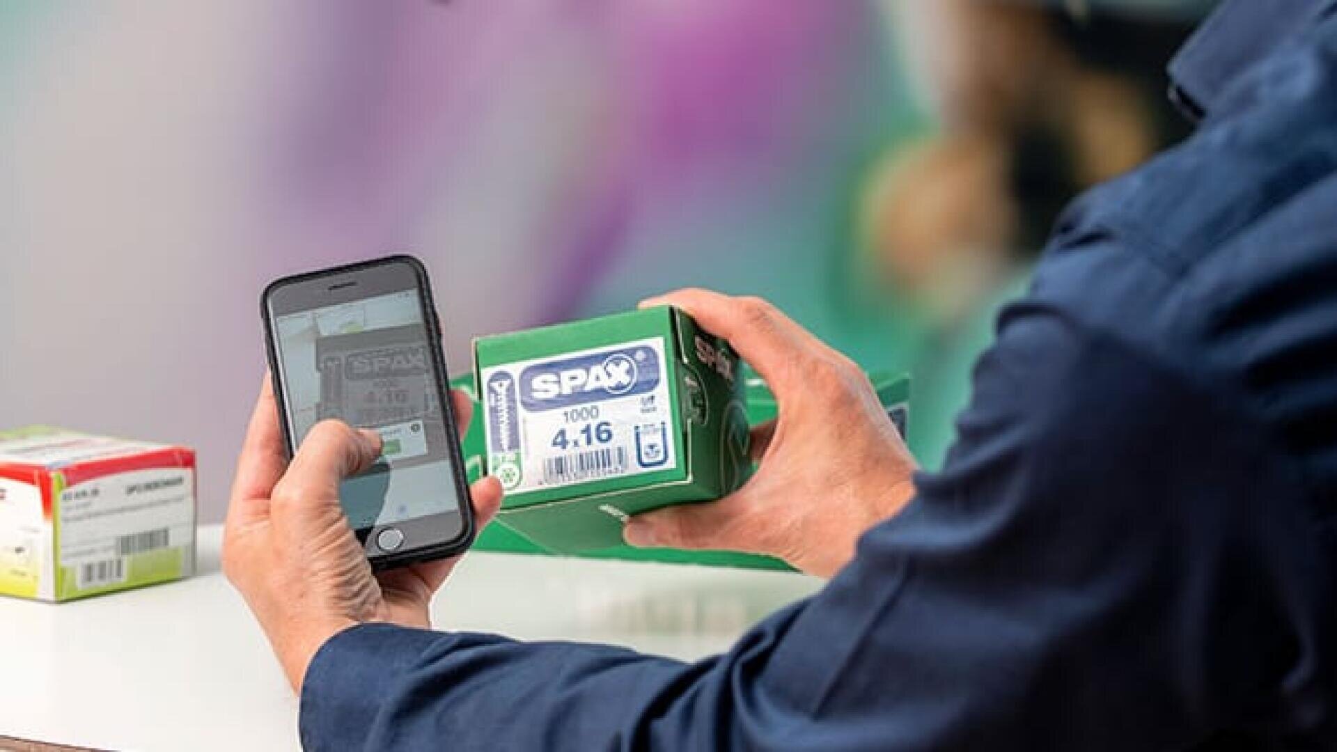 Eine Person scannt eine grüne SPAX-Schraubendose mit einem Smartphone. Eine andere Schachtel liegt auf dem Tisch in der Nähe, der Hintergrund ist unscharf.