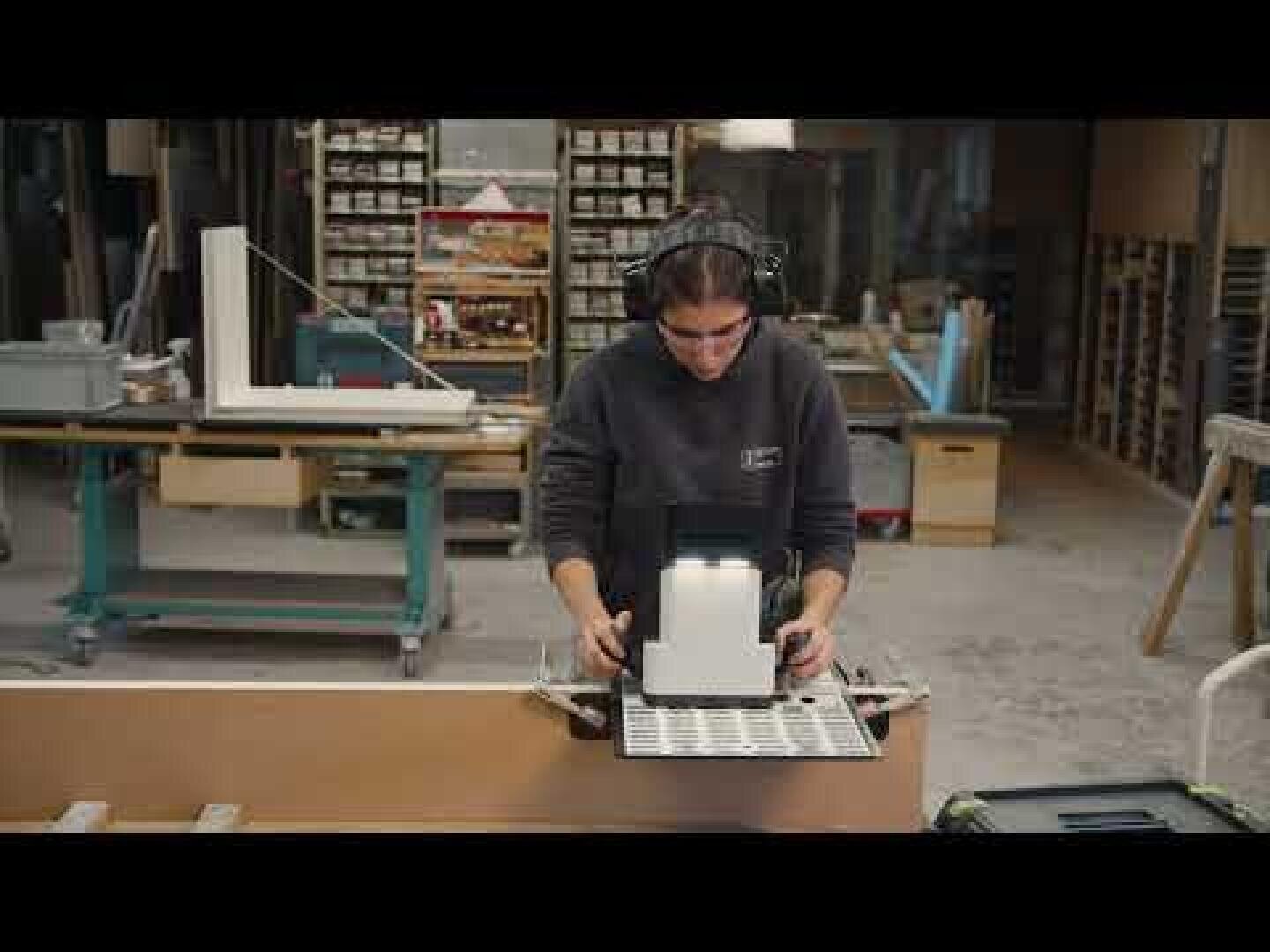 Eine Person mit Schutzbrille und Gehörschutz bedient eine Maschine in einer Holzwerkstatt, umgeben von Regalen mit Werkzeugen und Materialien.
