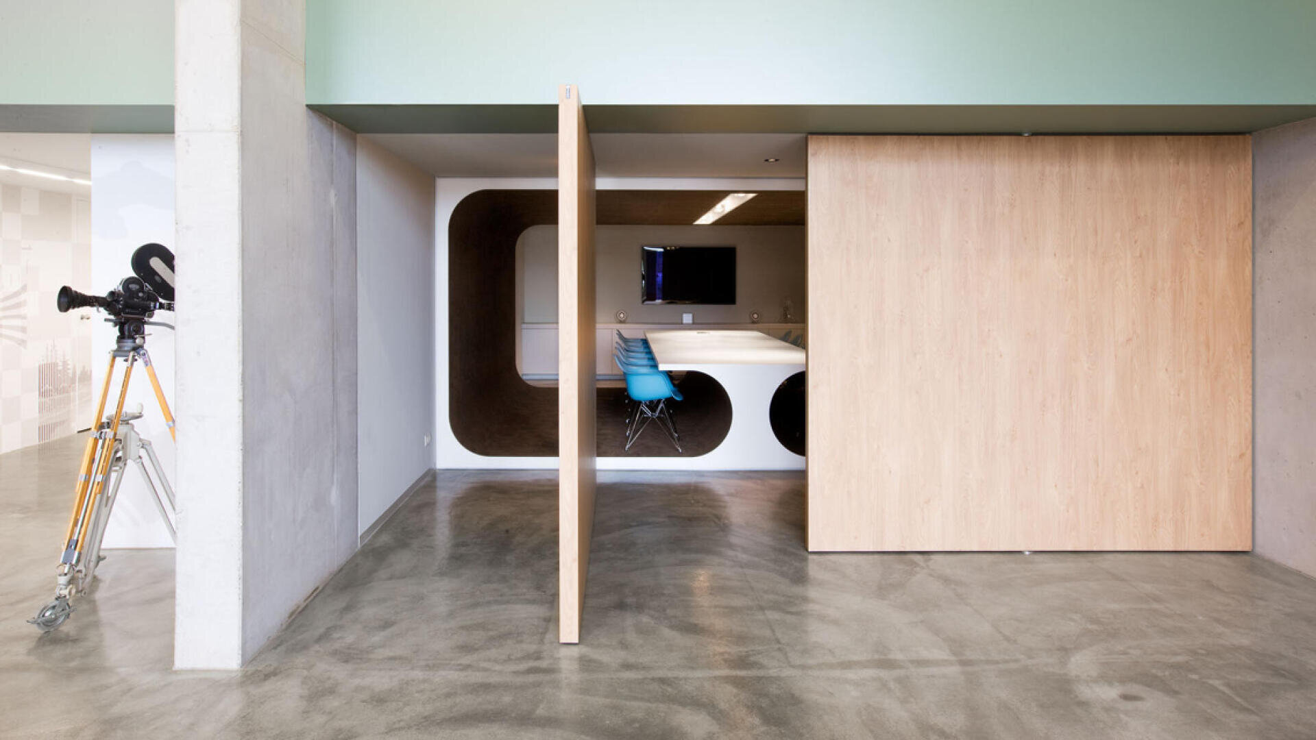 Un espace de bureau moderne avec des sols en béton poli, une porte coulissante en bois partiellement ouverte révélant une salle de conférence avec des chaises bleues, un moniteur fixé au mur et un vieil appareil photo sur trépied dans le couloir.