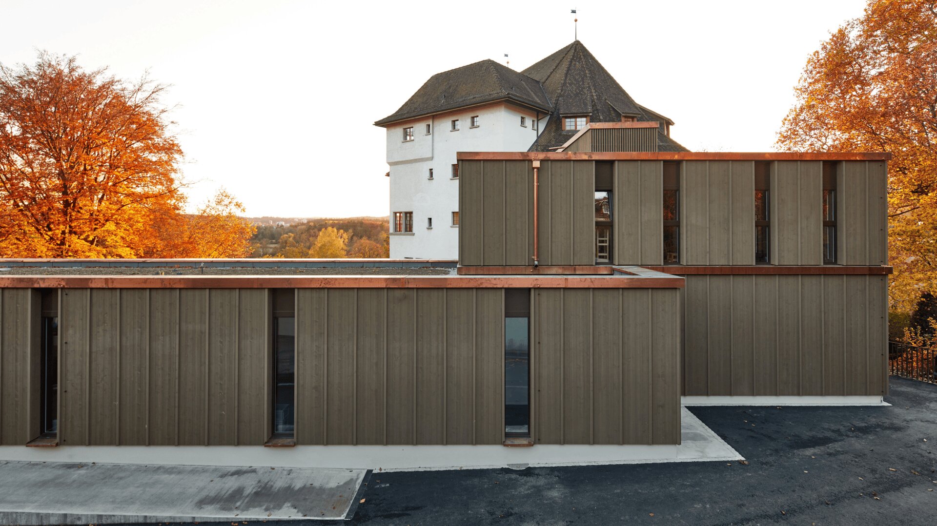 Ein modernes braunes Gebäude mit vertikaler Verkleidung steht vor einem historischen weißen Turm mit steilem Dach, umgeben von Herbstbäumen mit orangefarbenen Blättern.