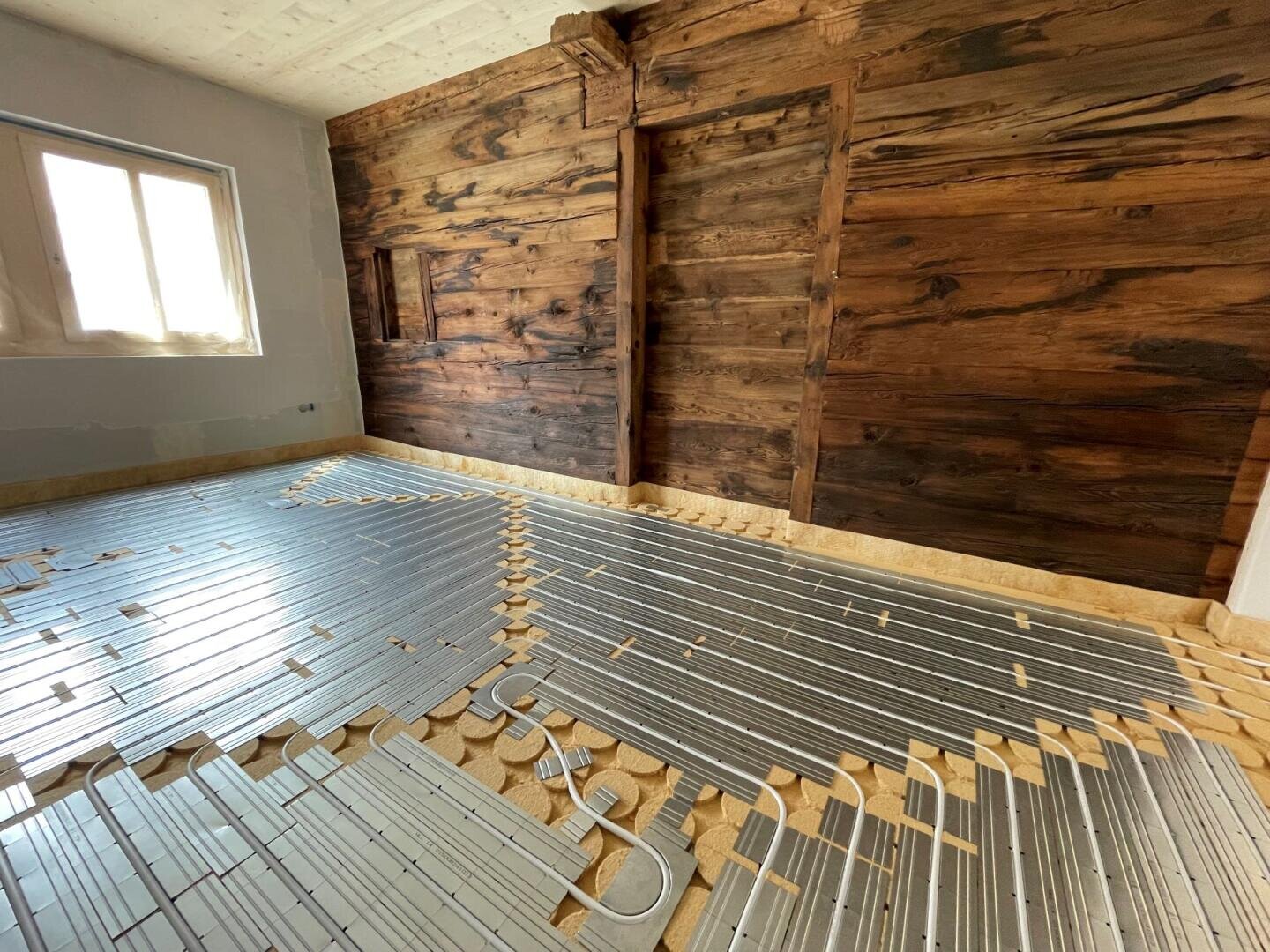 Pièce en cours de rénovation avec des panneaux métalliques de chauffage par le sol installés sur le sous-plancher, un mur d'accent en bois rustique, un plafond inachevé et la lumière du soleil passant par une fenêtre sur la gauche.