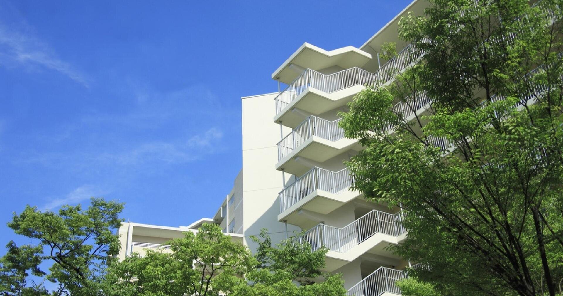 Un immeuble d'appartements moderne, avec ses balustrades blanches et ses balcons aux lignes angulaires, est partiellement dissimulé par des arbres verdoyants, sur fond de ciel bleu limpide.