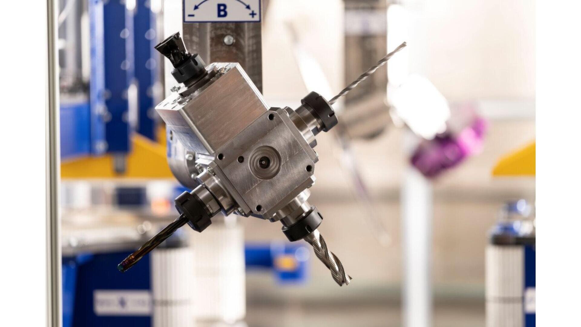 Gros plan d'une tête d'outil robotisée complexe avec de multiples composants métalliques, dont deux mèches différentes, fixée à une machine dans un environnement industriel moderne. L'arrière-plan montre des machines et des équipements flous.