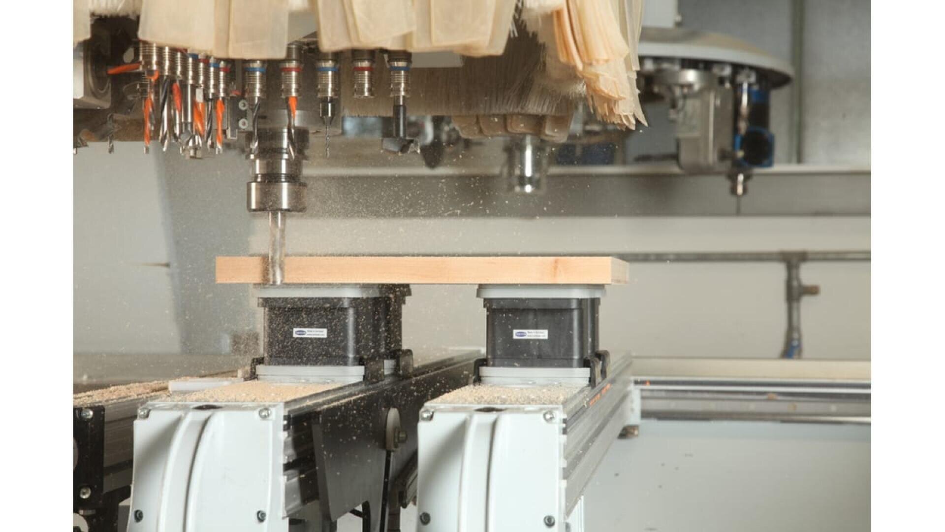 Une machine à commande numérique perce une pièce de bois maintenue par des pinces. Des copeaux de bois sont éparpillés et plusieurs mèches sont visibles au-dessus de la zone de travail.