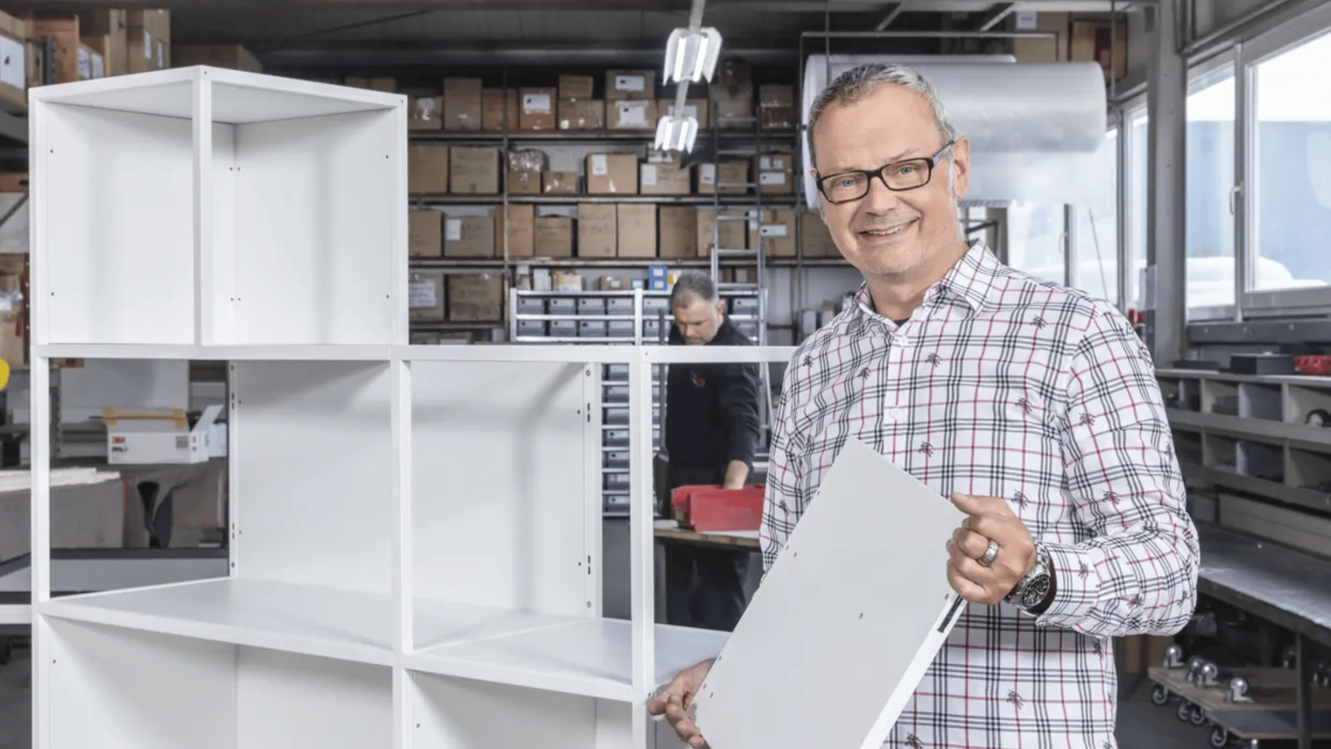 Ein lächelnder Mann mit Brille und kariertem Hemd hält eine weiße Platte, während er ein weißes Bücherregal in einer Werkstatt mit Regalen voller Kisten und einer weiteren Person im Hintergrund zusammenbaut.
