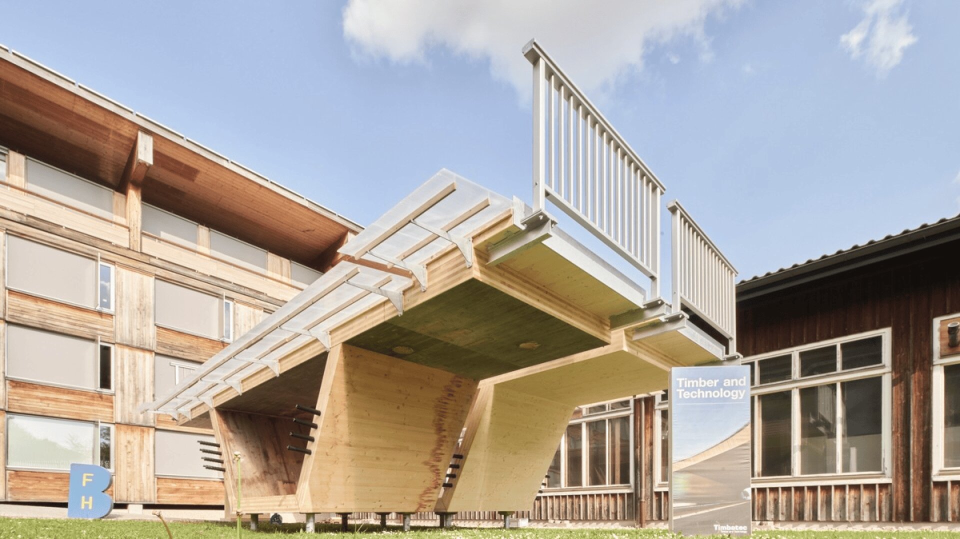 Eine moderne Holzkonstruktion mit einer auf dem Kopf stehenden Treppe steht im Freien zwischen zwei Holzgebäuden unter einem blauen Himmel mit Wolken. Auf einem Schild in der Nähe steht "Timber and Technology".