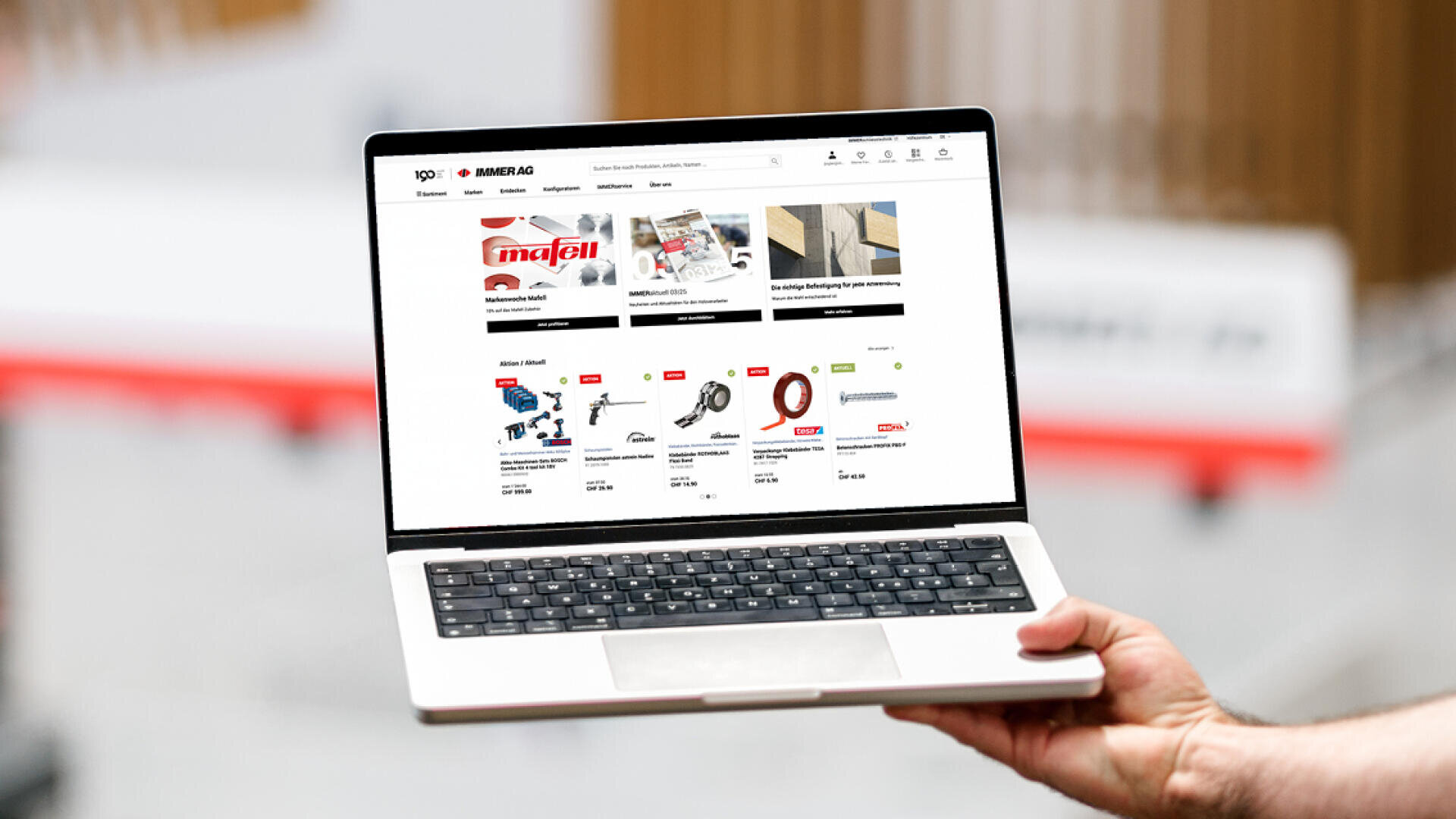 Une personne tient en main un ordinateur portable sur lequel s'affiche le site web d'une boutique en ligne proposant à la vente divers outils et appareils, dont des scies et des outils électriques. La mise en page du site présente des images de produits, des prix et des noms de marques.