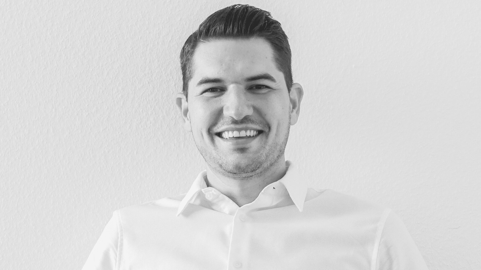 Homme souriant aux cheveux foncés, portant une chemise claire, photographié sur un fond clair et uni. L'image est en noir et blanc.