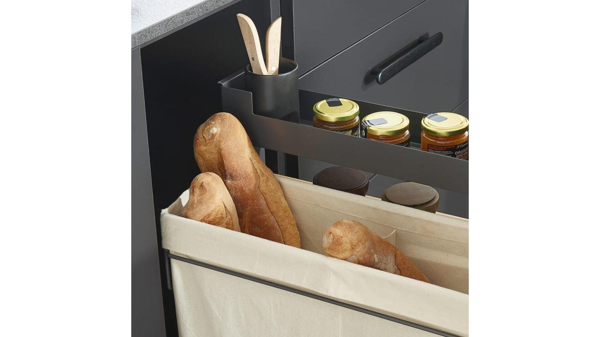Gros plan sur un organisateur de tiroirs de cuisine avec trois baguettes dans un bac en tissu beige, des bocaux de conserves sur une étagère et des ustensiles de cuisine dans un petit récipient noir.