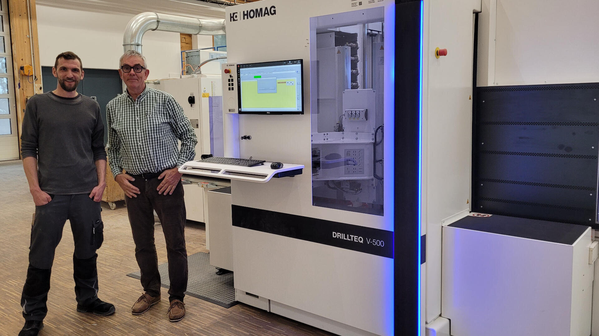 Zwei Männer stehen lächelnd neben einer großen HOMAG DRILLTEQ V-500 Industriemaschine in einer Werkstatt. Über einer an der Maschine angebrachten Tastatur zeigt ein Monitor Software an. Im Hintergrund sind weitere Geräte und Holzböden zu sehen.