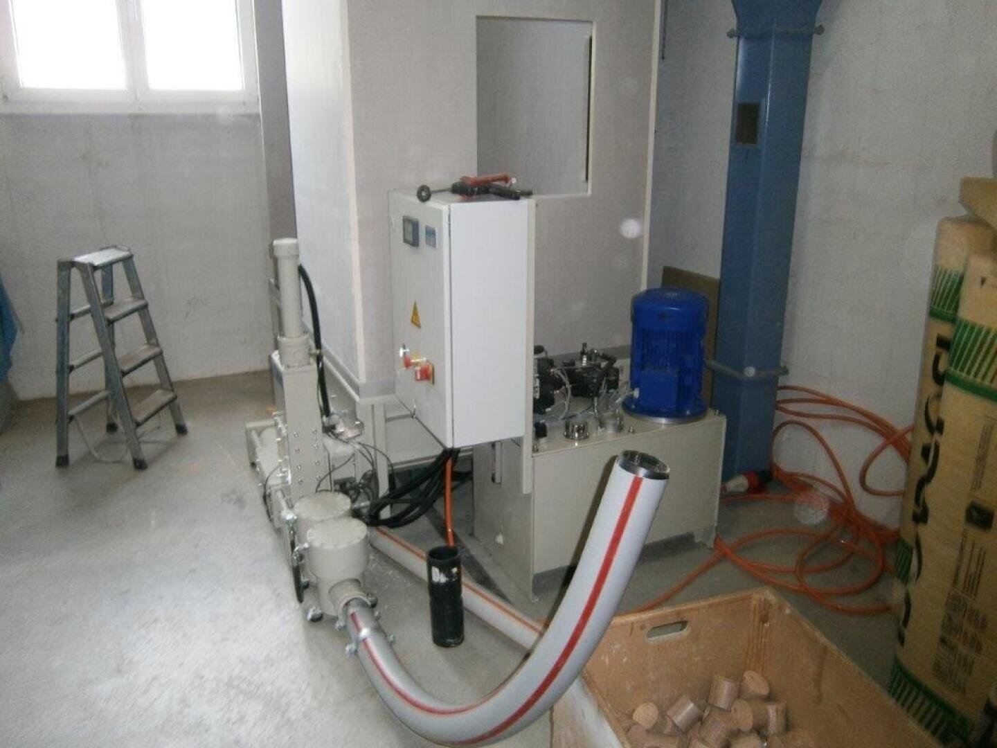 Ein Hydraulikaggregat mit Schalttafel, Schläuchen und einem blauen Motor ist in einem Industrieraum aufgestellt. Eine Stufenleiter, ein Karton mit zylindrischen Pellets und orangefarbene Kabel sind ebenfalls zu sehen.