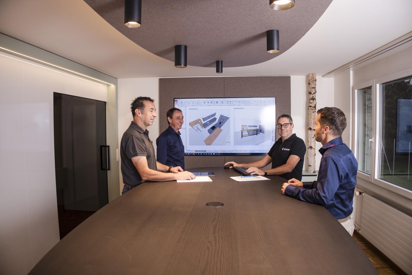 Vier Männer stehen um einen Konferenztisch in einem modernen Büro und diskutieren über architektonische Grundrisse, die auf einem großen Bildschirm hinter ihnen angezeigt werden. Einige von ihnen halten Papiere in der Hand und lächeln. Große Fenster sorgen für natürliches Licht.