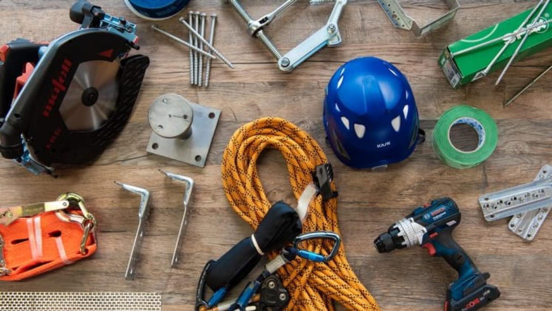 Verschiedene Bauwerkzeuge und Sicherheitsausrüstungen, darunter eine Bohrmaschine, ein blauer Schutzhelm, ein orangefarbenes Seil, eine Kreissäge, ein Klettergurt, grünes Klebeband, Metallhalterungen und Schrauben, die auf einer Holzfläche ausgebreitet sind.
