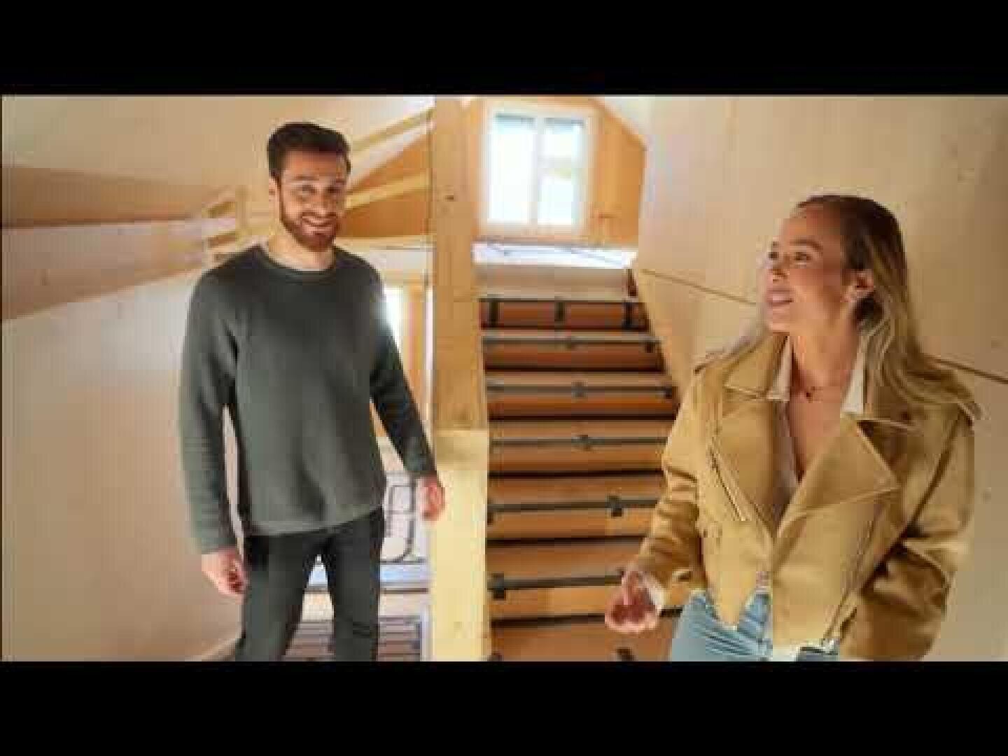 Ein Mann und eine Frau stehen in einer modernen Inneneinrichtung aus Holz in der Nähe einer Treppe. Der Mann trägt einen dunklen Pullover und die Frau eine beigefarbene Jacke und eine helle Jeans. Durch die Fenster im Hintergrund fällt Sonnenlicht ein.