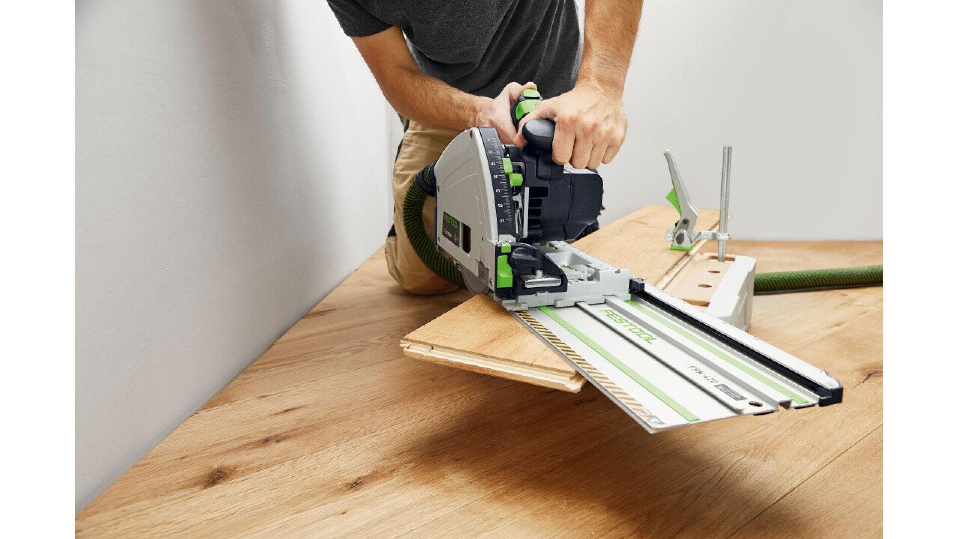 Une personne utilise une scie circulaire électrique avec un rail de guidage pour découper des planches de bois sur un plancher en bois dur, les mains tenant fermement l'outil pour plus de précision.
