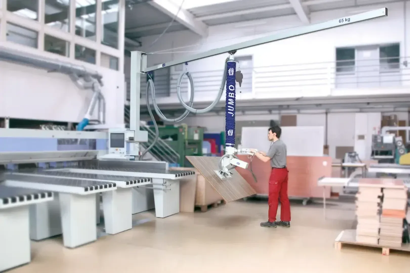Ein Arbeiter in einer roten Hose bedient einen mechanischen Hebearm mit einem Vakuumgreifer, um eine große Holzplatte in einer hellen, modernen Industrieanlage zu bewegen.