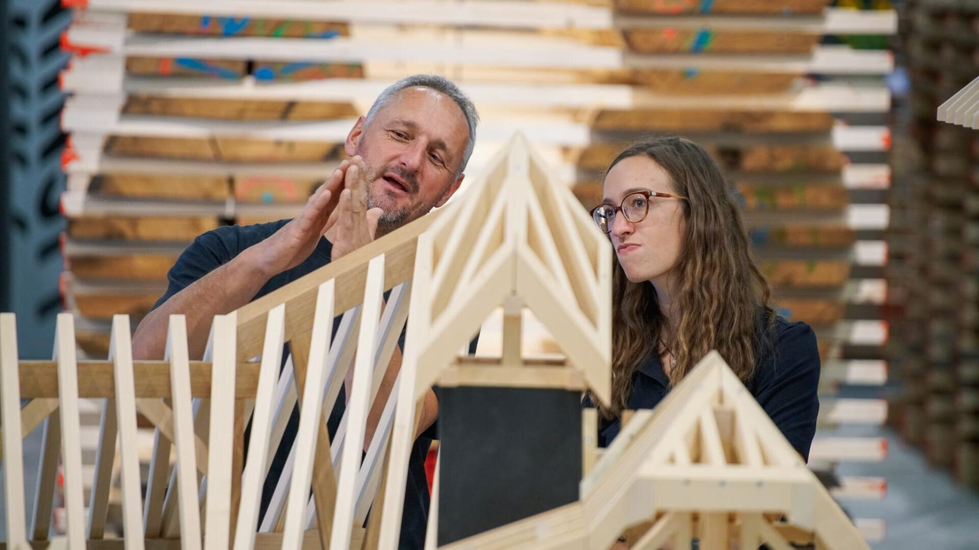Ein Mann erklärt einer jungen Frau mit Brille ein hölzernes Architekturmodell, wobei er mit seinen Händen gestikuliert, während sie aufmerksam zuhört. Im Hintergrund sind Stapel von ähnlichen Holzmodellen zu sehen.