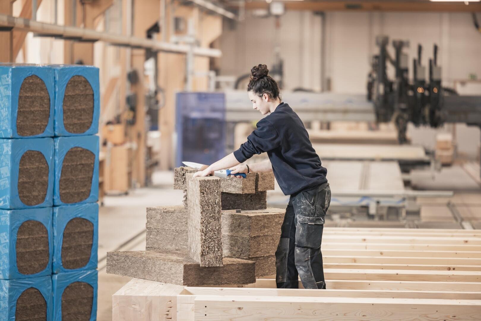 Eine Person in Arbeitskleidung stapelt große braune Dämmplatten in einer geräumigen, gut beleuchteten Holzwerkstatt mit Holzbalken, blauen Materialpaketen und verschiedenen Tischlergeräten im Hintergrund.
