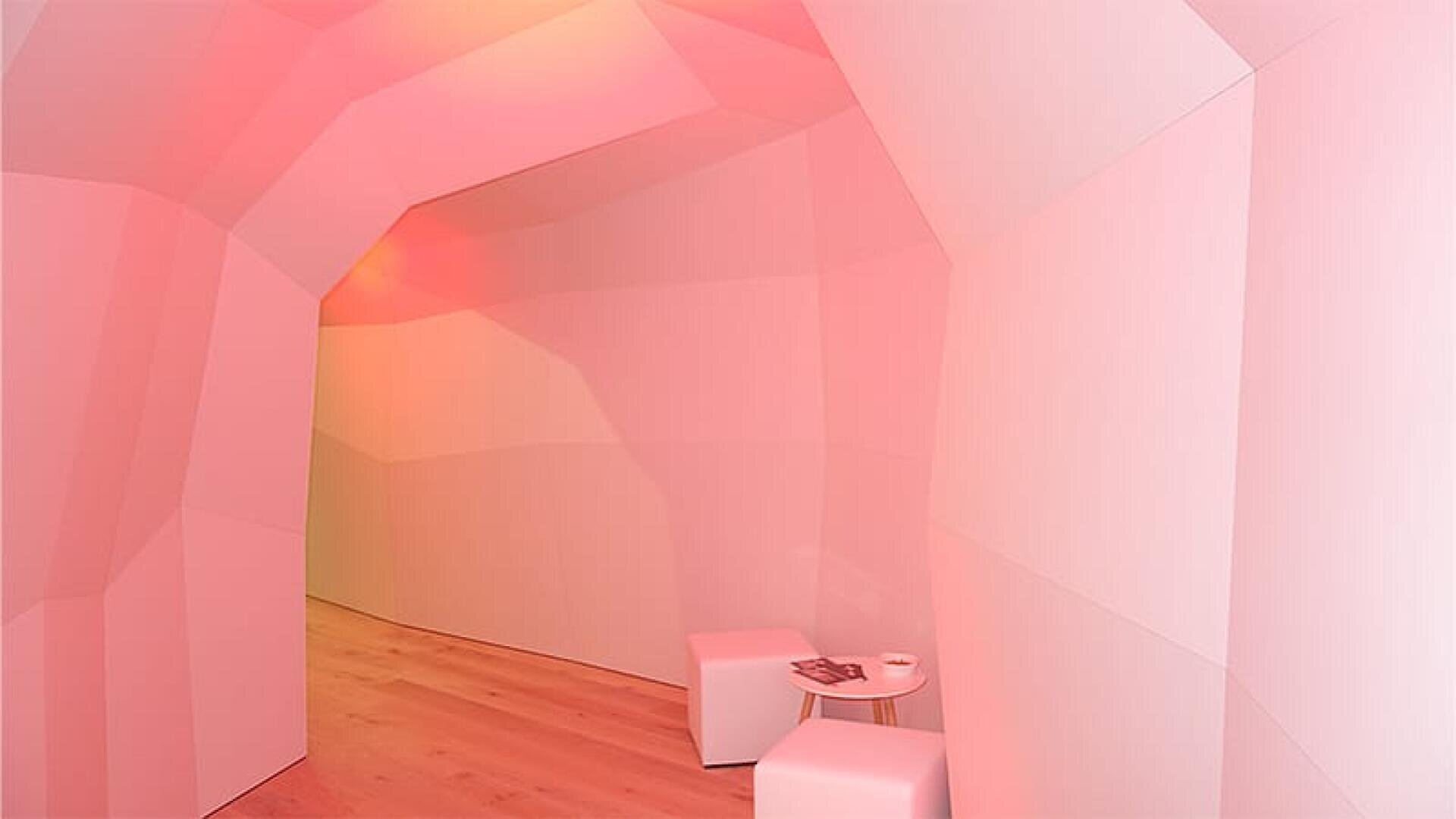 Une pièce aux murs géométriques à facettes, éclairée par une lumière rose, avec un sol en bois clair, deux sièges en cube blanc et une petite table ronde avec des magazines.