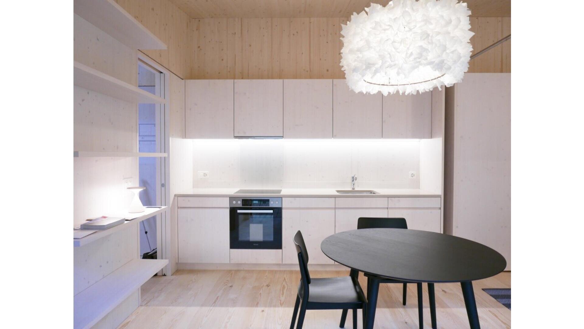 Cuisine moderne minimaliste avec des armoires blanches, des sols et des murs en bois clair, une table ronde noire avec deux chaises noires, des étagères ouvertes sur la gauche et une grande lampe suspendue à plumes blanches au-dessus de la table.
