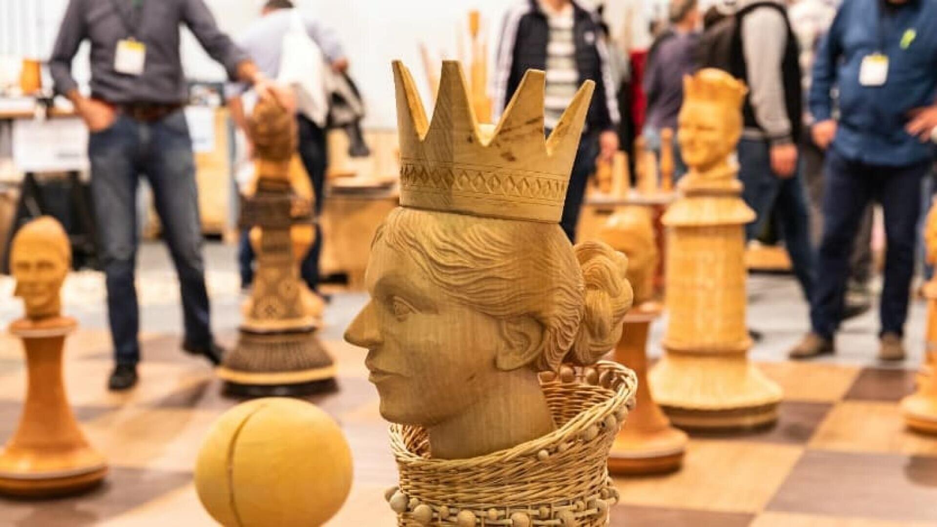 Eine große, kunstvoll geschnitzte hölzerne Schachfigur, die einer Königin ähnelt, ist auf einem riesigen Schachbrett ausgestellt, mit mehreren anderen übergroßen Holzfiguren und Menschen im Hintergrund.