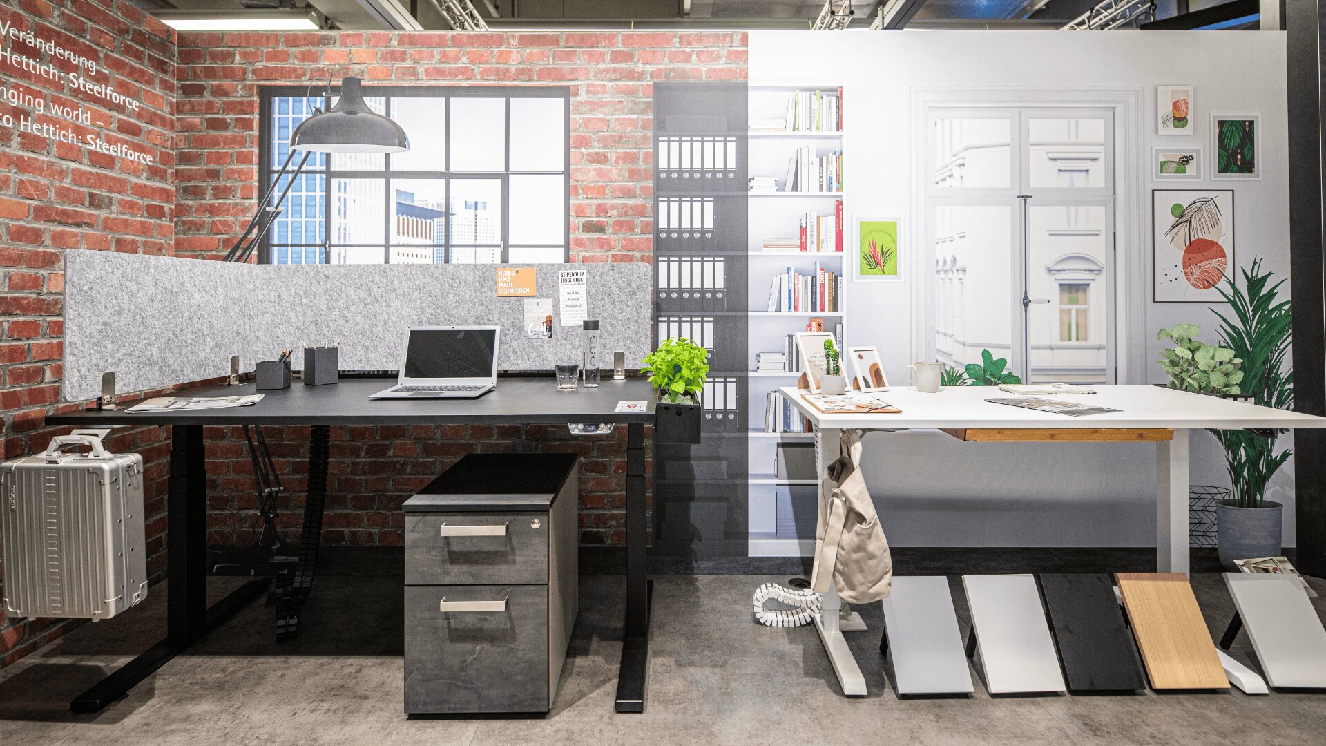 Moderner Büroarbeitsplatz mit zwei Schreibtischen, einem Laptop, einer Topfpflanze, einer Schreibtischlampe, Regalen mit Büchern, Mustertafeln und einer Backsteinwand im Hintergrund. Kunstdrucke und Dekoration sorgen für eine kreative, organisierte Atmosphäre.
