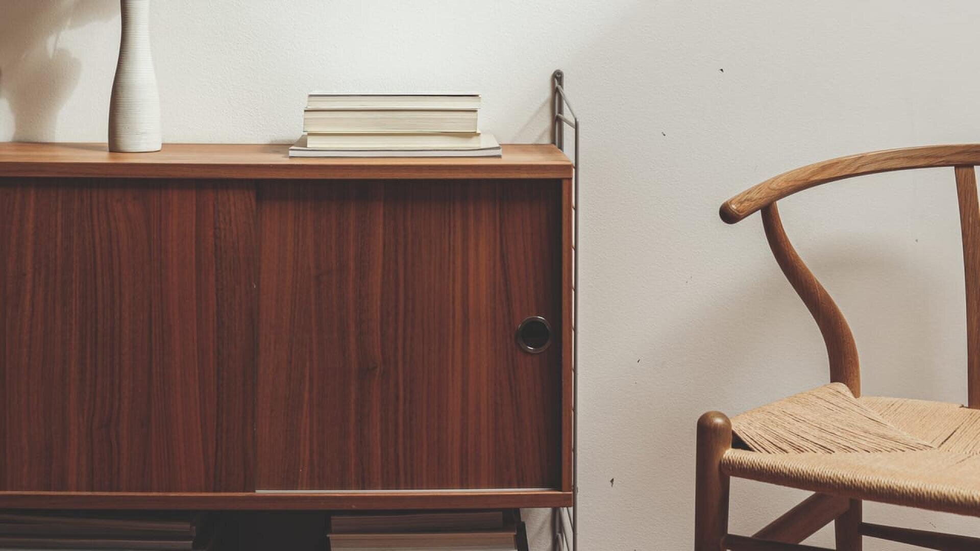 Un meuble en bois avec un vase blanc et des livres empilés se trouve à côté d'une chaise en bois avec une assise en tissu, contre un mur blanc dans un intérieur minimal et moderne.