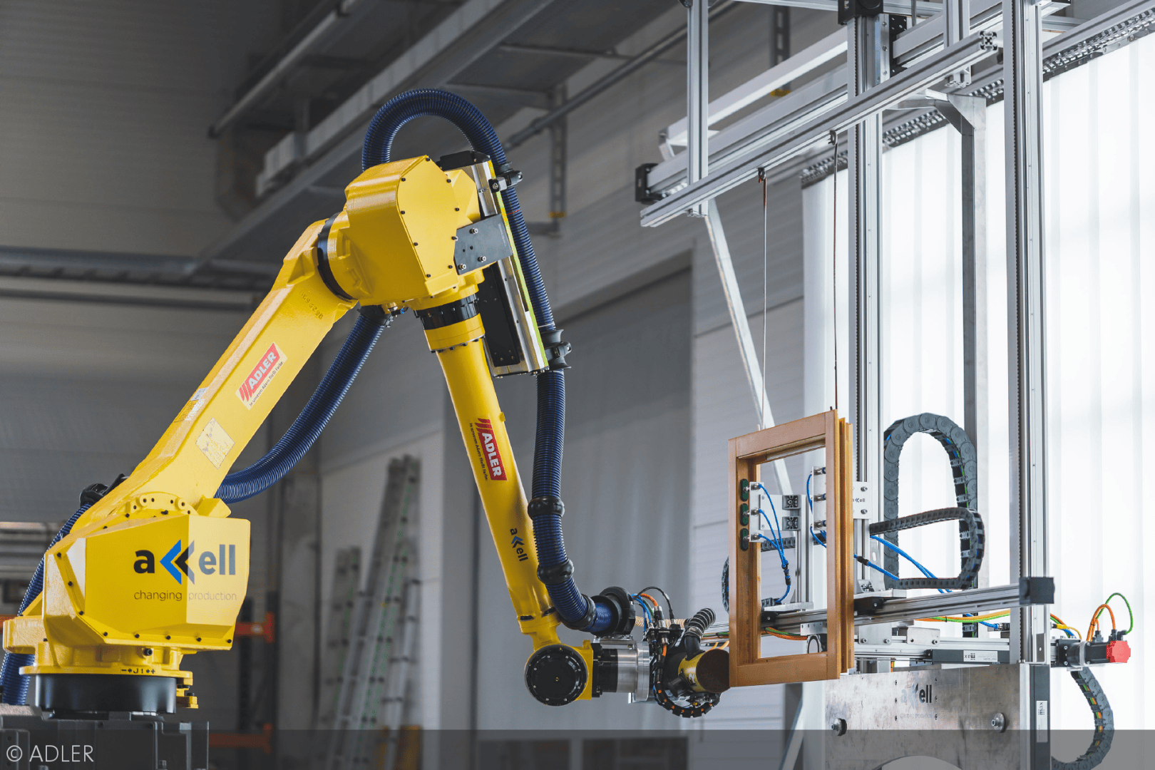 Ein gelber Industrieroboterarm montiert in einer modernen Fabrikumgebung einen Fensterrahmen. Der Arm ist mit Axell und KUKA beschriftet, im Hintergrund sind Maschinen und Kabel zu sehen.