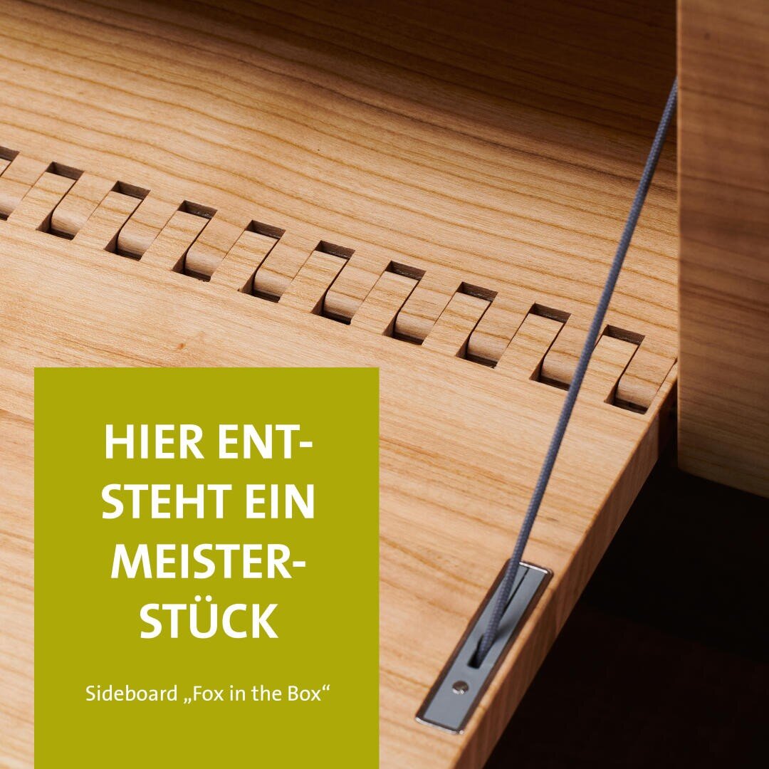 Nahaufnahme eines hölzernen Sideboards mit komplizierten Tischlerarbeiten und einem Metallkabel; deutscher Text auf einem grünen Rechteck: HIER ENTSTEHT EIN MEISTERSTÜCK. Sideboard Fox in the Box.