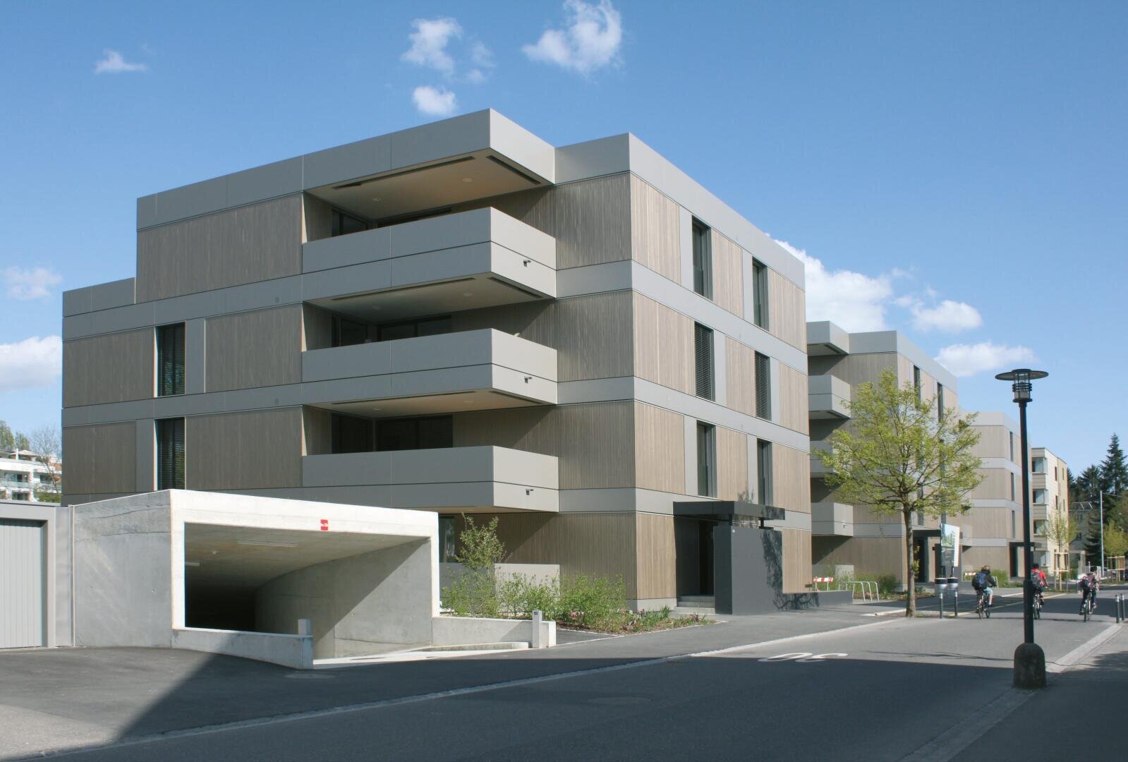 Modernes Mehrfamilienhaus mit klaren Linien und beigen Paneelen, mit großen Balkonen. Auf der linken Seite ist eine Garageneinfahrt zu sehen, und mehrere Personen gehen bei klarem, teilweise bewölktem Himmel die Straße entlang.