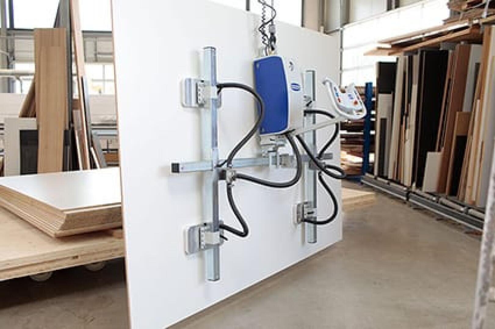 Eine große weiße Platte wird in einer Werkstatt durch eine Metallmaschine mit Kabeln und Klammern vertikal befestigt, umgeben von Stapeln aus Holz und Baumaterialien.