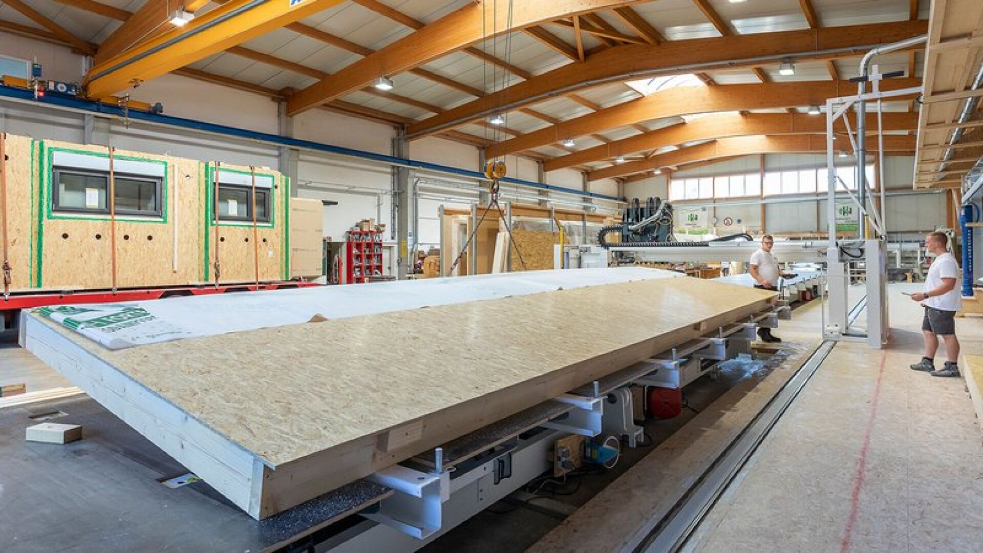 Zwei Arbeiter montieren in einer geräumigen, gut beleuchteten Fabrik eine große vorgefertigte Holzwandplatte. In der Anlage sind Holzbalken, Maschinen und eine weitere teilweise fertige Wand im Hintergrund zu sehen.