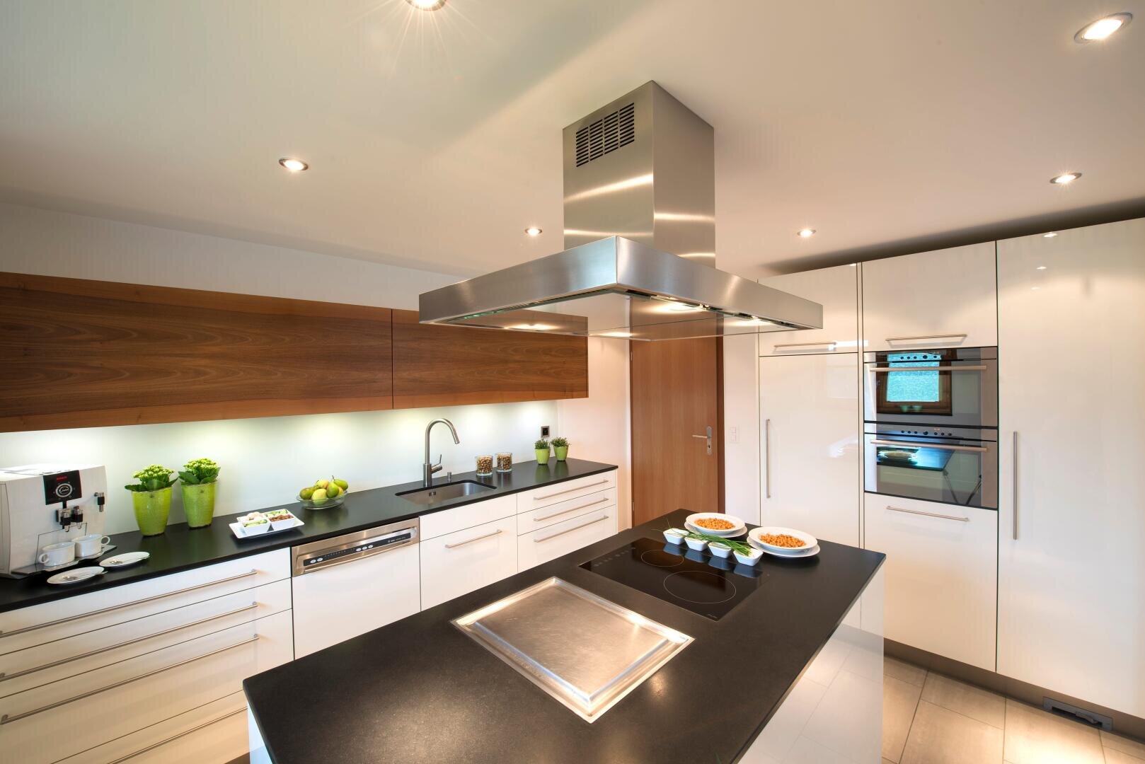 Moderne Küche mit weißen Schränken, Holzakzenten, einer Insel mit schwarzer Arbeitsplatte, eingebautem Backofen, Elektroherd, Dunstabzugshaube aus rostfreiem Stahl und verschiedenem Geschirr und Utensilien in ordentlicher Anordnung.