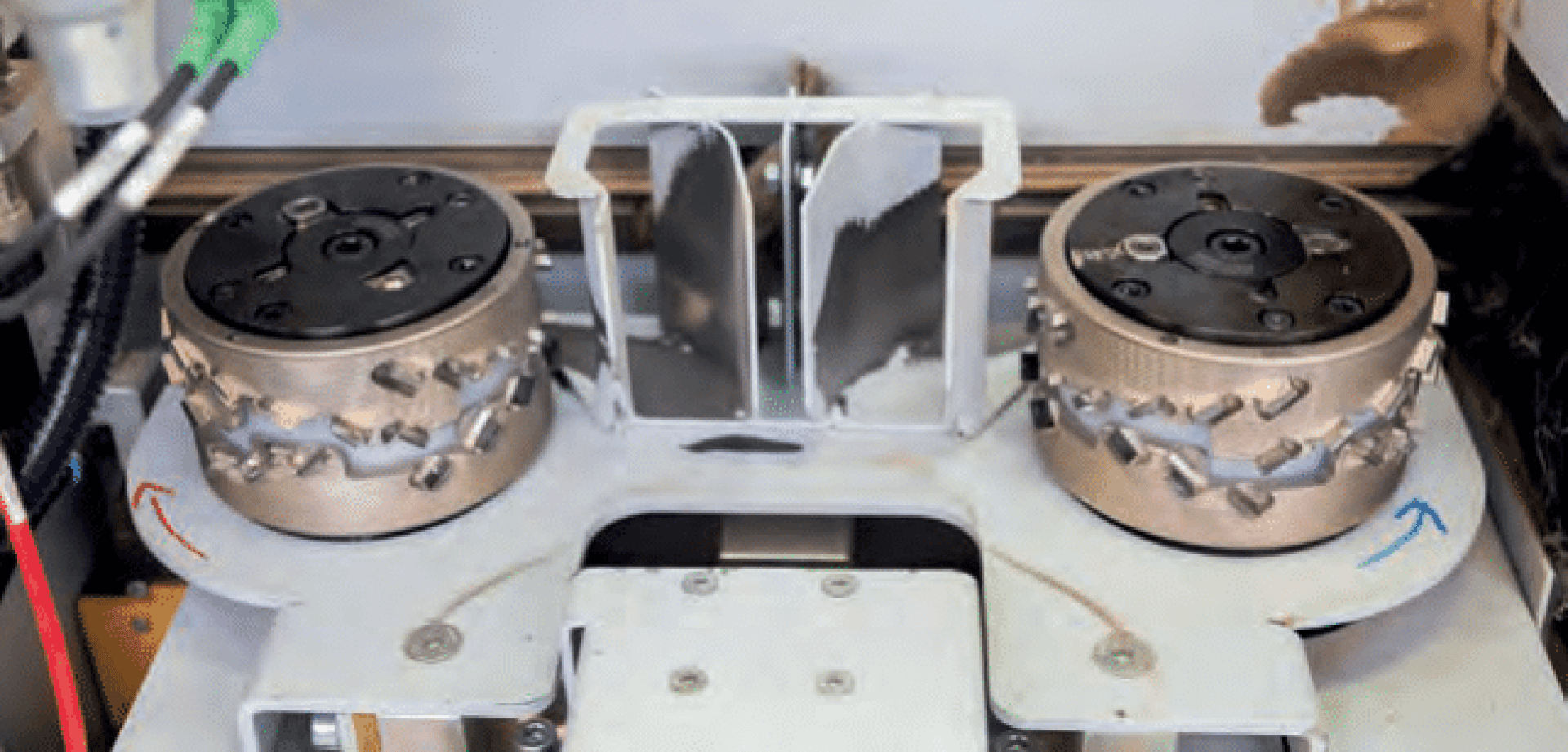 Nahaufnahme eines industriellen Maschinenteils mit zwei rotierenden Metallschleifscheiben mit Schneidkanten, die nebeneinander angeordnet sind und einem Metallteiler in der Mitte gegenüberstehen. Die Pfeile zeigen die Drehrichtung an.