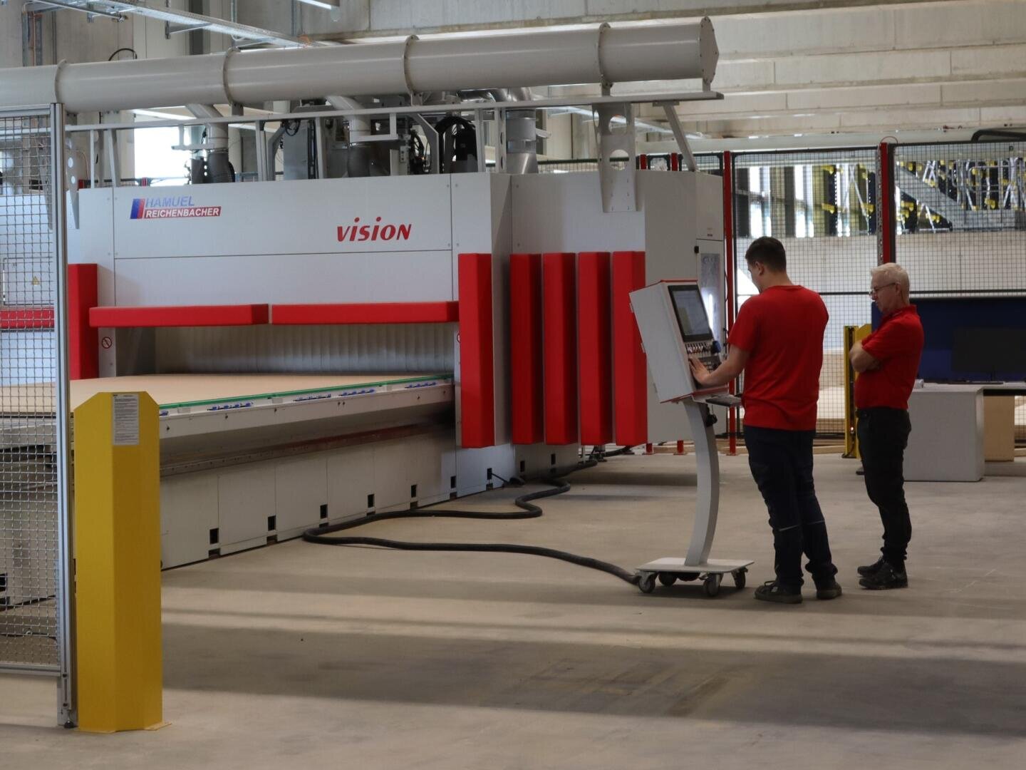 Dans une usine spacieuse et moderne, deux hommes en chemise rouge font fonctionner une grande machine industrielle appelée VISION. L'un d'eux utilise un terminal d'ordinateur tandis que l'autre se tient à proximité, en train d'observer.