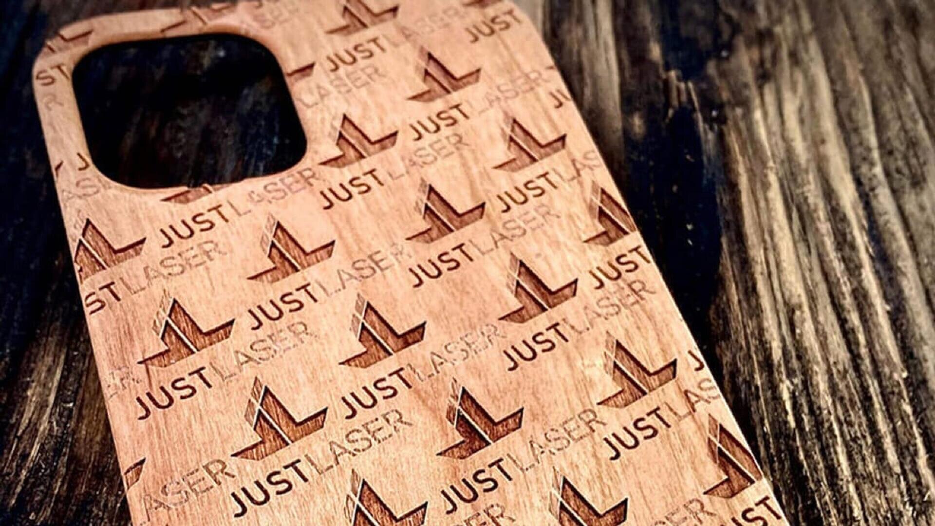 Un étui de téléphone en bois gravé d'un motif répété des mots JUST LASER et d'un logo JL stylisé, reposant sur une surface en bois foncé.
