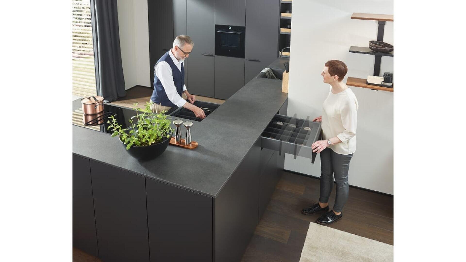 Un homme et une femme se trouvent dans une cuisine moderne aux meubles sombres. La femme ouvre un tiroir, tandis que l'homme range des ustensiles près de l'évier. Un grand bol noir avec des plantes est posé sur l'îlot de cuisine.