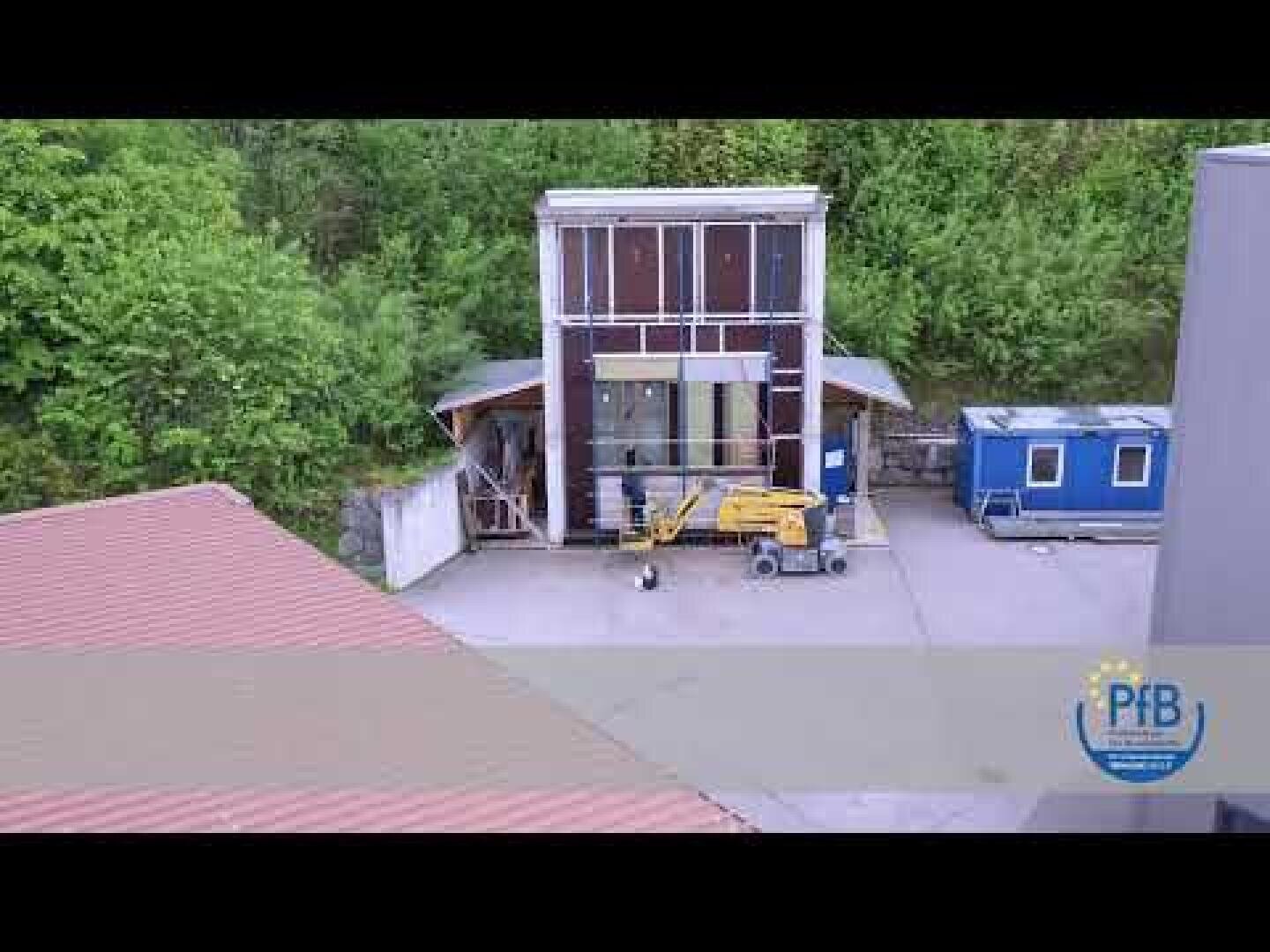 Ein kleines Industriegebäude mit großen Fenstern steht neben einer blauen tragbaren Hütte. Eine gelbe Gondel ist im Freien geparkt, umgeben von Bäumen und anderen Strukturen. Das PFB-Logo ist in der Ecke zu sehen.