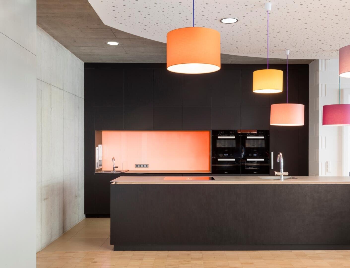 Cuisine moderne avec des armoires noires, un rebord de couleur pêche, deux fours, un îlot avec un évier et plusieurs lampes suspendues colorées suspendues à un plafond blanc à motifs.