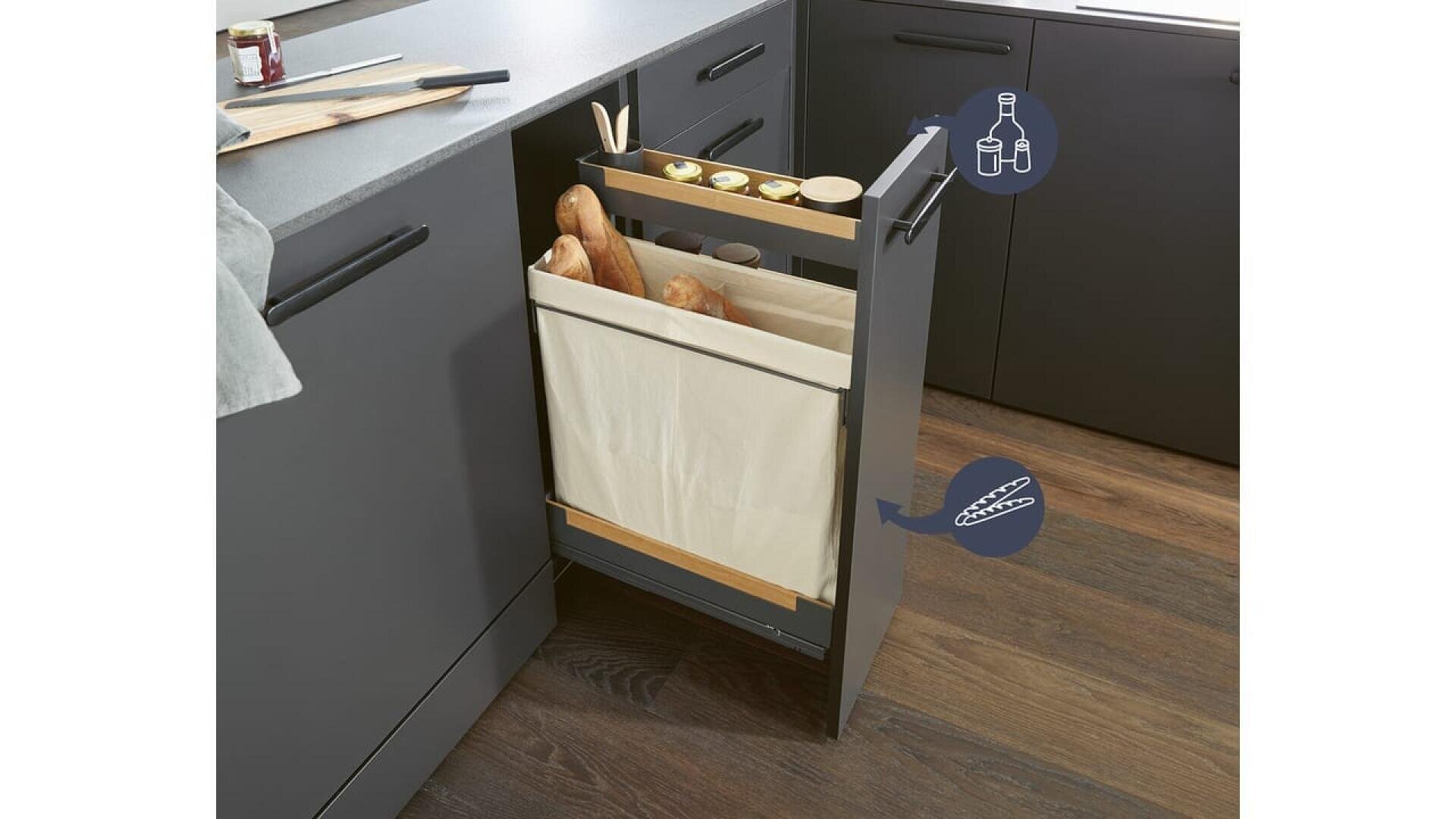 Une cuisine moderne présente un meuble coulissant avec un bac de rangement en tissu beige contenant des baguettes et des ustensiles de cuisine. Le meuble est encastré dans une armoire foncée et étiqueté avec des icônes pour les bouteilles et le pain.