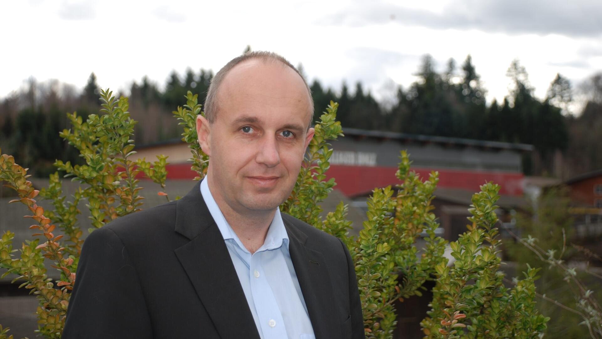 Ein Mann in einer Anzugjacke und einem hellblauen Hemd steht draußen vor grünen Büschen, Bäumen und einem verschwommenen Gebäude im Hintergrund unter einem wolkenverhangenen Himmel.