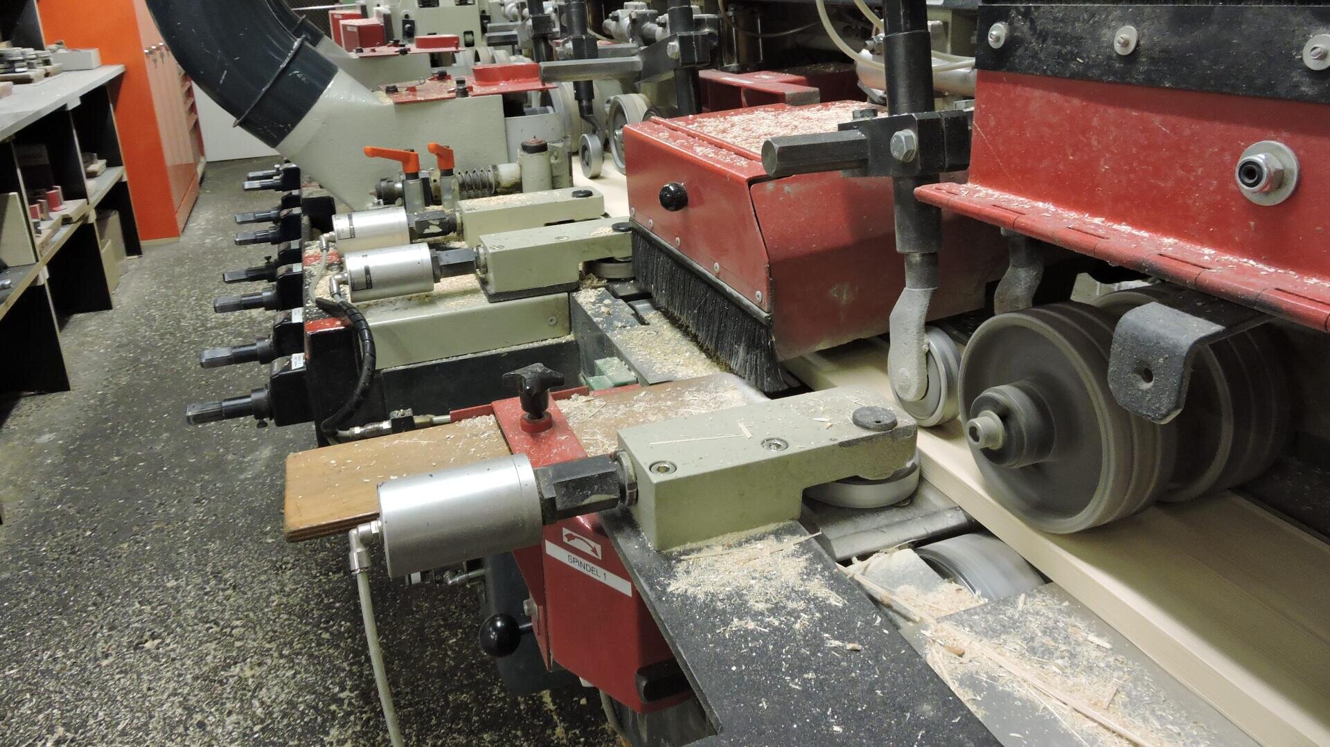 Nahaufnahme von Holzbearbeitungsmaschinen in einer Fabrik. Zu sehen sind mehrere rote und graue Maschinen, die Holzbretter bearbeiten, mit Sägemehl auf dem Boden und den Geräten.