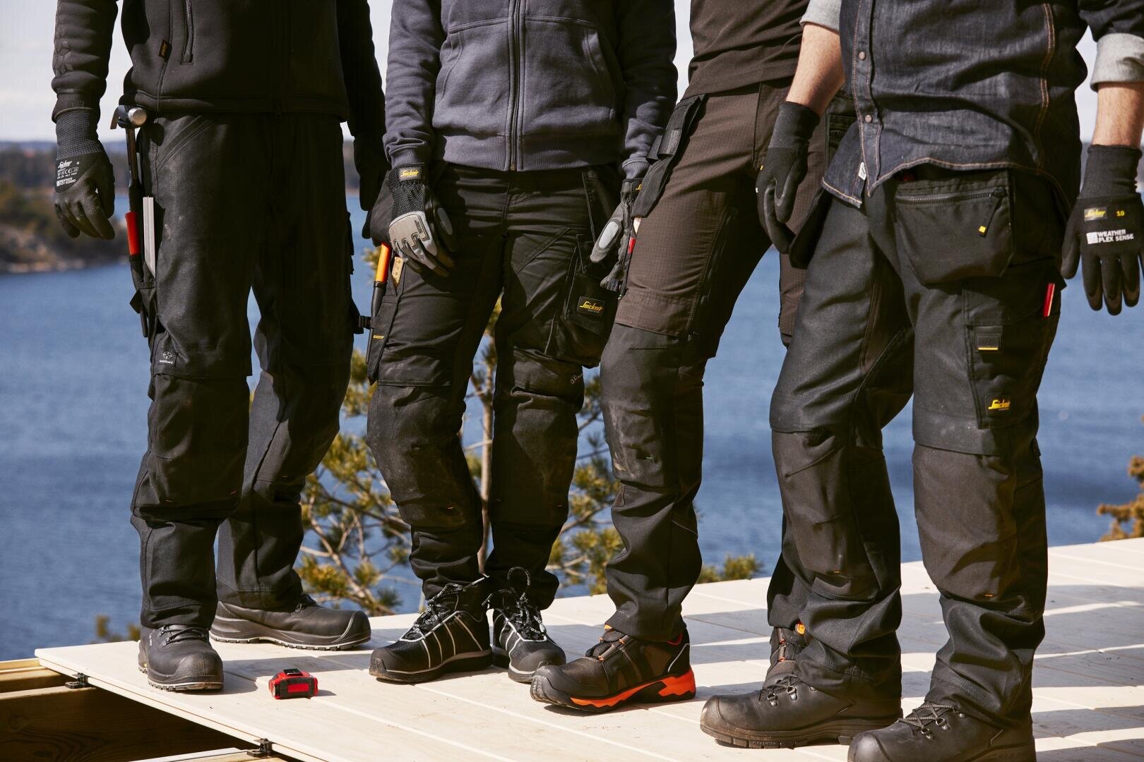 Quatre personnes, vêtues de vêtements de travail et de gants, se tiennent debout sur une plateforme en bois, à l'extérieur, près d'un point d'eau. Des outils sont attachés à leurs pantalons et elles portent des chaussures robustes, suggérant qu'elles travaillent dans la construction ou la menuiserie.