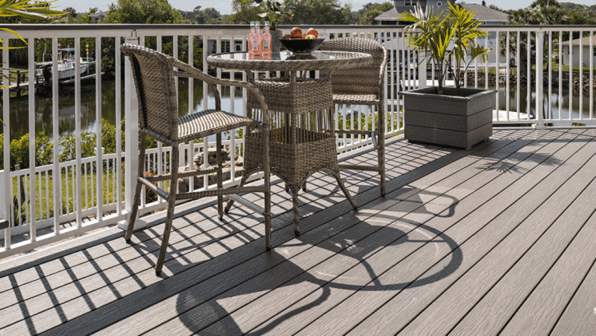 Eine sonnenbeschienene Terrasse mit zwei Korbstühlen und einem runden Tisch mit Obst und Getränken, mit Blick auf Wasser und Bäume, mit Geländern, die Schatten auf den Holzboden werfen.