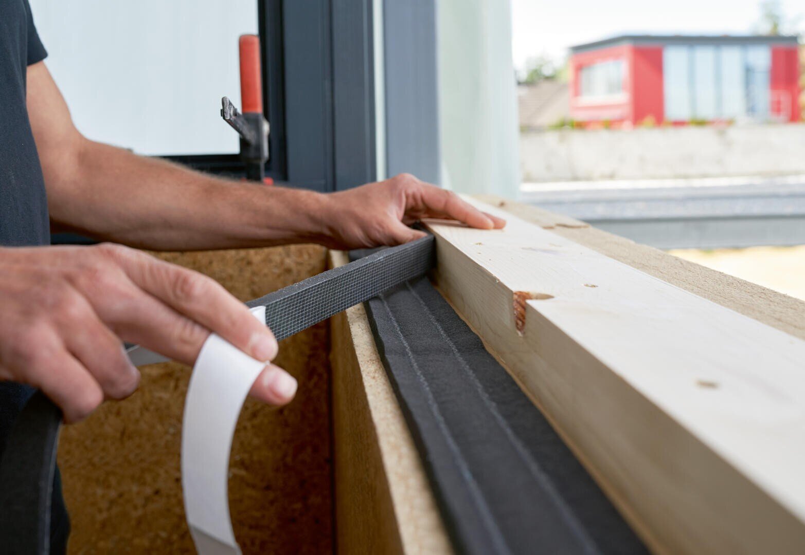 Eine Person klebt Klebeband auf einen Holzbalken in der Nähe eines Fensters, möglicherweise als Vorbereitung für die Isolierung oder Abdichtung bei Bau- oder Renovierungsarbeiten.