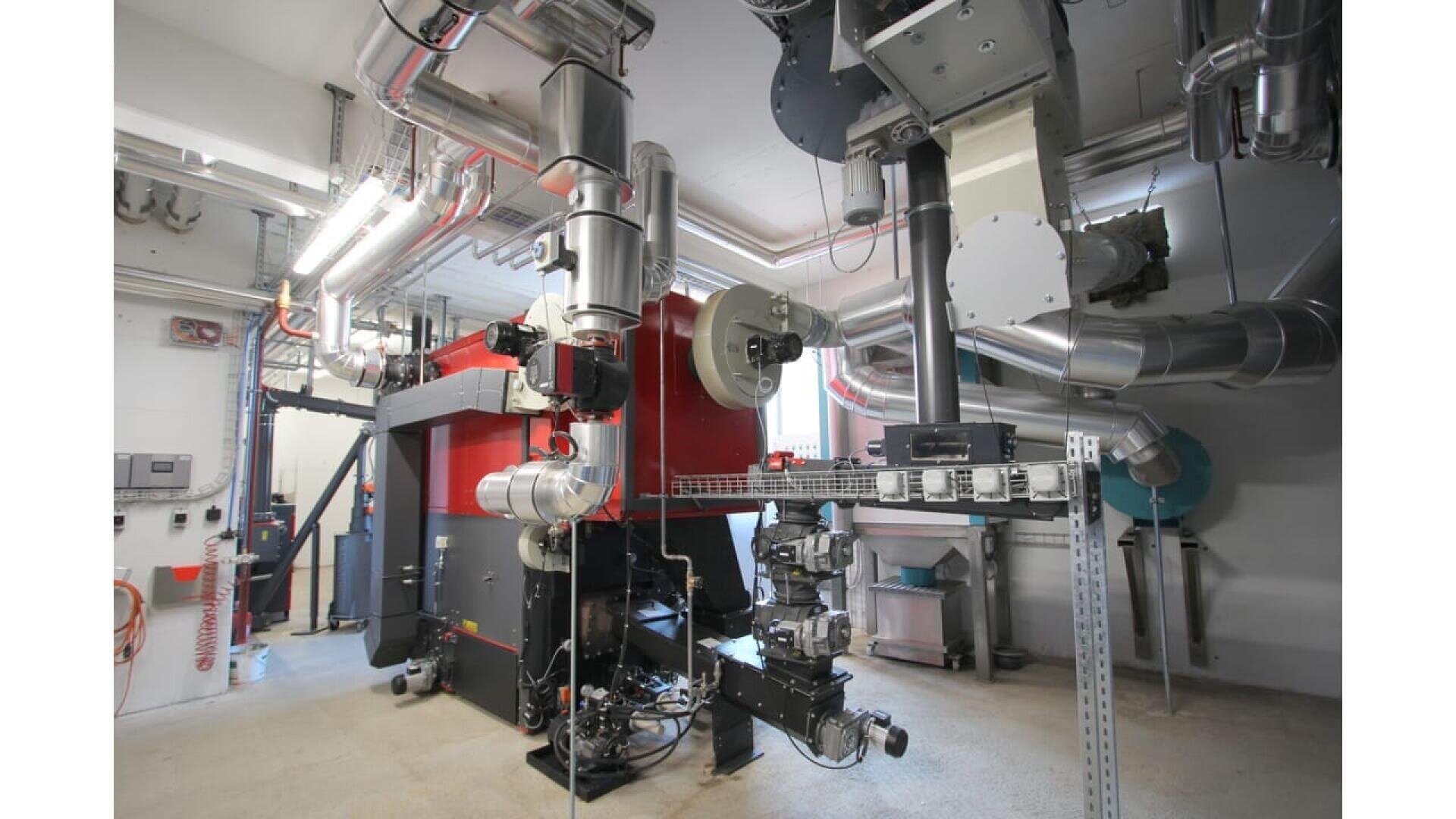 Un grand système de chaudière industrielle avec une unité principale rouge et noire, entourée de nombreux tuyaux métalliques, de vannes et de composants électriques dans une salle de service propre et bien éclairée.
