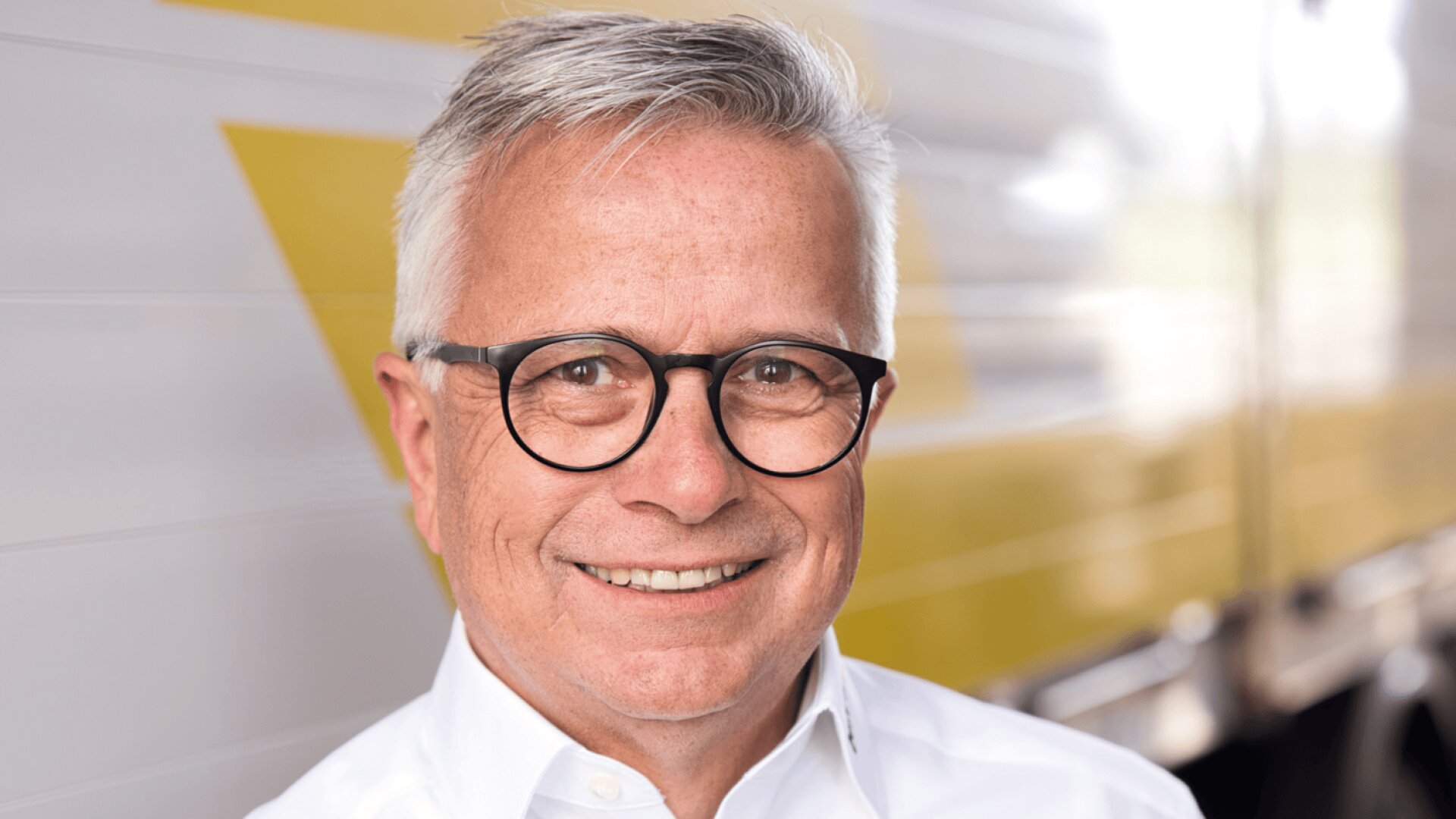 Un homme souriant, âgé, aux cheveux gris courts, portant des lunettes et une chemise blanche se tient devant un arrière-plan flou avec des lignes diagonales jaunes et blanches.